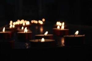 A row of tealight candles glowing softly in a dimly lit environment, creating a warm and serene atmosphere. The candles' flickering flames are reflected on the dark surface below.