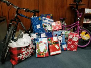 A collection of wrapped holiday gifts is displayed on the floor, including two bicycles—one blue and one pink. The wrapped presents feature festive designs with snowflakes, Santa Claus, and holiday greetings.