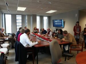 A group of people sitting around a long conference table in an office setting, engaged in conversation and eating. A decorative centerpiece adorns the table, and a screen on the wall displays a presentation. Large windows let in natural light.