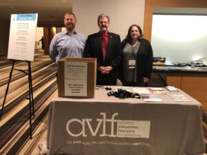 Three individuals stand behind a table with a tablecloth displaying the 