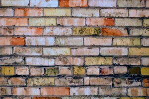 A close-up view of a weathered brick wall, showing a variety of colors from orange and yellow to brown and gray, with uneven textures and mortar in between. The bricks are arranged in a horizontal, alternating pattern.