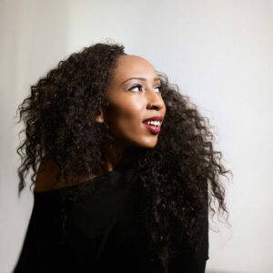 A woman with long, curly hair and wearing a black off-shoulder top looks upward, smiling. The background is softly lit, adding a warm glow to the scene.