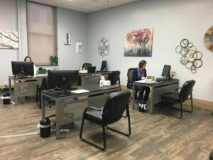 A modern office with two people working at separate desks. Each desk has a computer, paperwork, and office supplies. The room has light-colored walls, abstract art on the walls, and wood flooring. The office appears organized and tidy.
