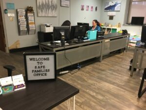 An office with two people working at computers behind a long desk. A sign in the foreground reads 