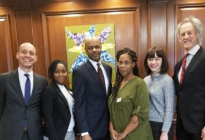 A group of six people, three men and three women, stand smiling in front of a wooden paneled wall. A colorful abstract painting is displayed behind them. They are all dressed in business attire.