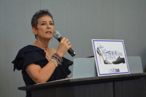 A person with short hair speaks into a microphone at a podium. They wear a dark outfit and bracelets. A sign on the podium reads 