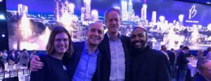 Four people smiling in front of a large backdrop featuring a cityscape with bright lights. They appear to be at a formal event, as tables with place settings and a crowd are visible in the background.