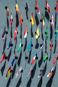 A vertical wall displays multiple international flags in neat rows, casting long shadows against a light blue background. The flags include various colors and patterns, representing different countries.