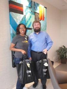 Two people standing and smiling indoors, each holding black bags with a logo. The woman is wearing a gray T-shirt, and the man is in a blue shirt. A colorful abstract painting is in the background.