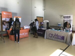 People visiting booths at a community health fair in a gymnasium. The WellCare and AVLF tables display information and staff engage with attendees. Signs read 