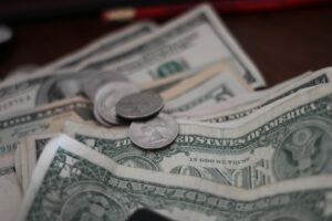 A close-up of several U.S. dollar bills, including one-dollar notes, with a few coins scattered on top. The bills and coins are laid out on a table, suggesting currency counting or a financial concept.