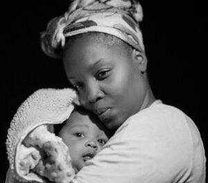 A black and white photo of a woman holding a baby wrapped in a blanket. The woman wears a headscarf and is looking at the camera with a gentle expression. The baby peeks out from the blanket, also gazing at the camera.