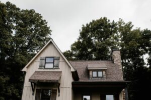 A two-story house with a peaked roof and dormer windows, surrounded by tall green trees. The exterior is a mix of stone and wood paneling, with a visibly overcast sky above.