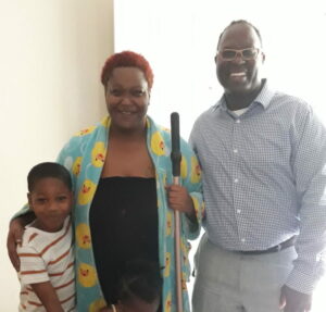 A smiling group of three people standing indoors. A woman in a duck-patterned robe holds a cane, with a young boy hugging her. A man stands next to them wearing glasses and a checkered shirt.