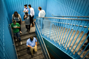 People walking up and down a vibrant blue staircase inside a building. Some are using their phones, while others engage in conversation. The railing and walls are painted in uniform blue stripes, adding a dynamic feel to the scene.