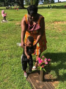 A woman and a young child stand together at a gravesite. The woman holds the child as they look at a headstone adorned with pink flowers. The setting is a grassy cemetery with other headstones and floral arrangements visible in the background.