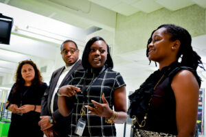 Four people are standing indoors, engaging in conversation. Three women and one man are facing each other, appearing attentive and engaged. They are dressed in professional attire. The background shows a hallway with fluorescent lighting.