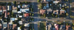 Aerial view of a suburban neighborhood with rows of houses, trees, and streets intersecting. The houses have various roof colors and patterns, and the area appears well-organized with a clear layout.