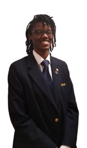 A person smiling, wearing a dark formal suit with gold buttons, a white shirt, and a dark tie. They have glasses and short braided hair. The background is white.
