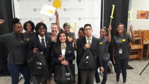 A group of smiling students and adults pose in front of a backdrop with 