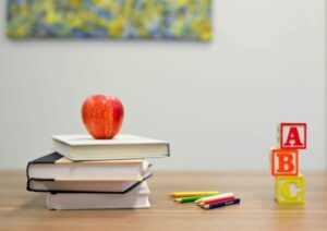 A stack of books with an apple on top sits on a wooden table. To the right, there are colorful alphabet blocks spelling 