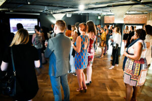 A diverse group of people stands attentively in a modern room with wooden flooring, facing a large screen displaying a presentation. Some hold drinks, and ambient lighting creates a warm atmosphere.
