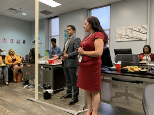A man and a woman stand in front of a group of seated people in an office setting. They are talking, and the woman is wearing a red dress. A table with food and drinks is visible, and people are listening attentively. Artwork adorns the walls.