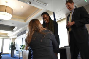 Three people are interacting in a conference room. A seated woman with long hair faces two standing individuals, a woman with braided hair and a man in a suit, who are smiling. A presentation screen and podium are in the background.