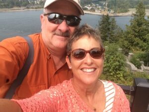 A man and a woman smiling and taking a selfie on a sunny day. The man is wearing a cap and orange shirt, and the woman is wearing sunglasses and a peach-colored top. They are standing near a body of water with trees and a bridge in the background.
