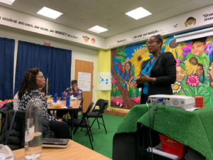 A woman stands speaking in a classroom decorated with a colorful mural. Two women sit at a table listening, with papers and a water bottle visible. A projector faces a screen, and motivational quotes adorn the walls.