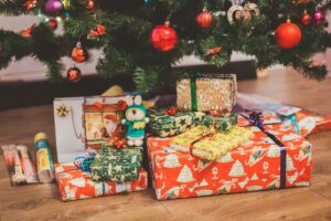 A variety of wrapped Christmas presents in colorful paper are neatly arranged under a decorated Christmas tree. The tree features ornaments in red, gold, and other vibrant colors. A plush toy is visible among the gifts on a wooden floor.