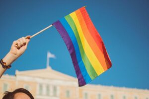 A hand holding a rainbow pride flag against a clear blue sky, with a historic building softly blurred in the background.