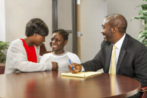 A woman and a child sit at a round table, warmly interacting with a man in a suit who holds a notepad and pen. They are indoors, with a plant in the background, suggesting a professional or school setting.