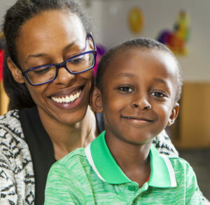A woman wearing glasses is smiling at a young boy in a green polo shirt. They are sitting close together, both looking happy. The background is softly blurred, suggesting an indoor setting.