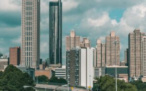 A cityscape featuring tall skyscrapers and modern high-rise buildings under a partly cloudy sky. Trees and urban infrastructure, including roads and signs, are visible in the foreground.