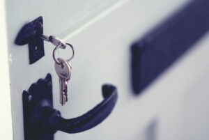 A close-up image of a door with a black handle, featuring two keys inserted into the keyhole. A name tag with an emblem is attached to the keyring. The door appears slightly ajar, with a bright, soft focus background.