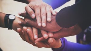 A diverse group of people place their hands on top of each other in a display of unity and teamwork. The hands vary in skin tones, and one person is wearing a black wristwatch. The background is blurred.
