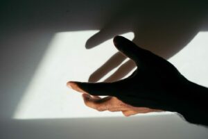 A hand reaching out is casting a shadow on a wall, both the hand and its shadow are partially illuminated by a soft light, creating a dramatic and intriguing visual effect. The background is a neutral color.