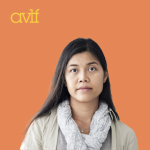 A woman with long dark hair, wearing a light-colored scarf and beige jacket, stands against an orange background. A logo with the letters 