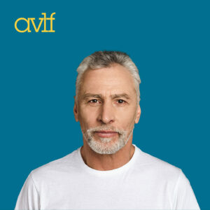 A man with short gray hair and a beard wearing a white t-shirt against a blue background with the letters 