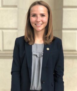 A person with shoulder-length brown hair is smiling, wearing a dark blazer over a patterned blouse. They stand in front of a light-colored, structured background.