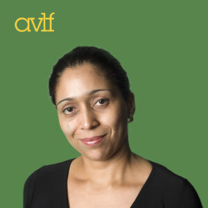 A person with a neutral expression is standing against a solid green background, wearing a black top. The yellow letters 