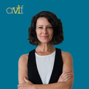 A woman with shoulder-length dark hair stands confidently against a teal background, wearing a white top and black vest. She has crossed arms and a slight smile. The letters 