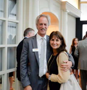 A man in a suit and tie stands with his arm around a woman wearing a dark top and cream cardigan. They are smiling at the camera in a room with other people in the background. The man wears a nametag.