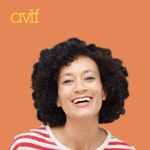 A person with curly hair smiles broadly while wearing a red and white striped shirt. The background is orange with the letters 