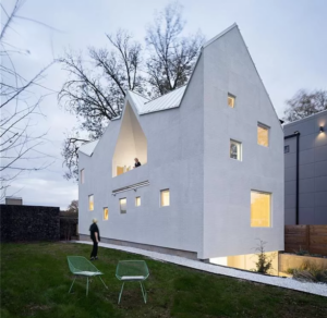 Modern white house with geometric design features triangular roofs and multiple windows. A man stands in the yard next to two green chairs. The setting is a grassy area with a paved path and trees in the background. Sky is overcast.