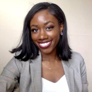 A person with shoulder-length black hair smiles warmly. They are wearing a plaid blazer over a white top. The background is neutral, adding focus to their expression.