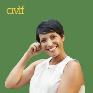 A person with short dark hair smiles warmly, wearing a sleeveless white top. They are sitting in front of a green background with the letters 