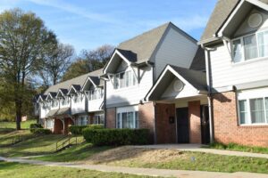 A row of residential townhouses with red brick lower walls and white upper siding. The homes feature gabled roofs and white-framed windows. Green lawns and bushes line the front, and the sky is clear and blue.