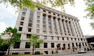 A large historic courthouse with tall columns and multiple stories. The building is surrounded by a few trees, and there are people walking along the sidewalk. The sky is partly cloudy.